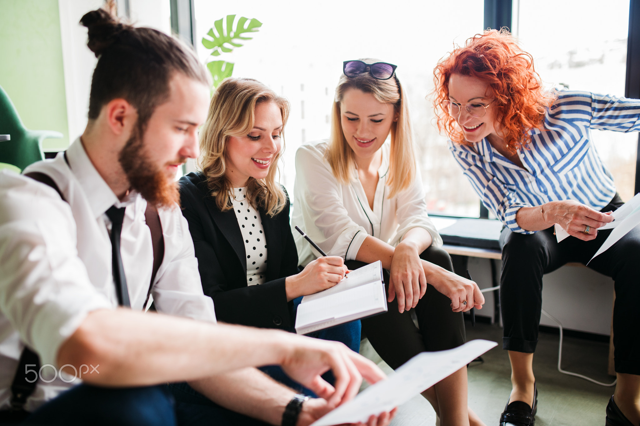 A group of young business people sitting on the floor in an office, talking.