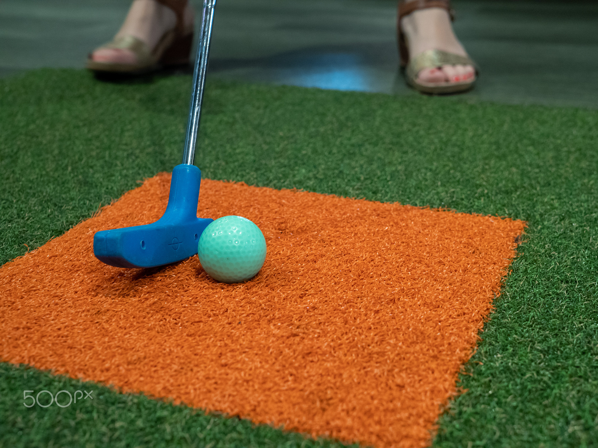 Blue putter on turf next to green golf ball on miniature golf course with woman hitting