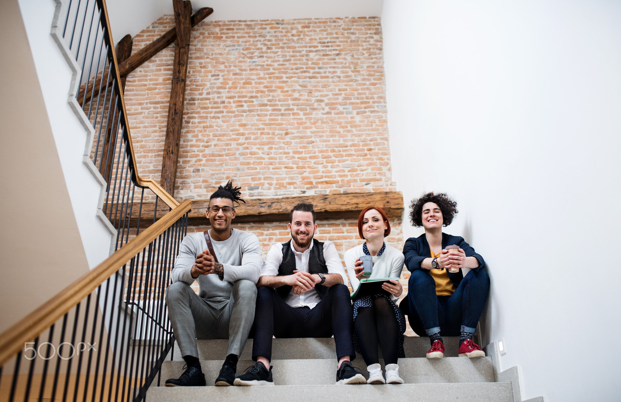 Group of young businesspeople sitting on stairs indoors, looking at camera.